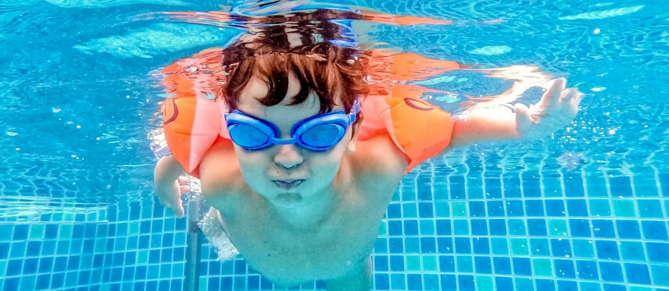 A child swimming underwater in a pool, wearing blue goggles and orange armbands, playfully holding their breath with a tiled pool floor visible in the background.