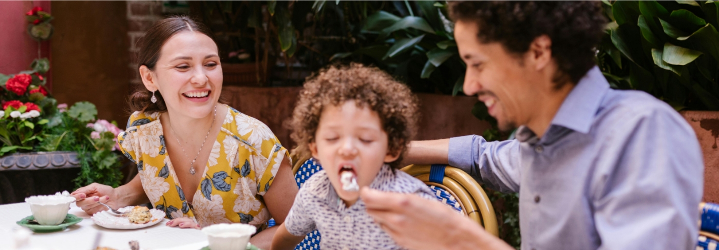 A happy family enjoying a meal together at an outdoor café, with a mother smiling, a father feeding their child a spoonful of dessert, and lush greenery and flowers in the background.