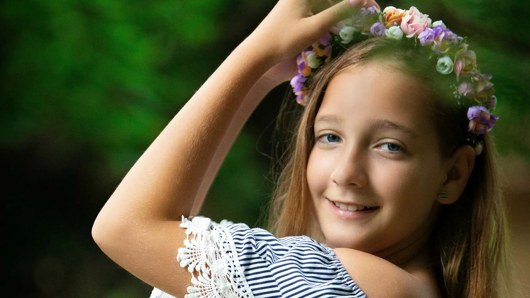 A young girl smiling and adjusting her floral crown, wearing a striped lace-trimmed top, with a blurred green forest background.