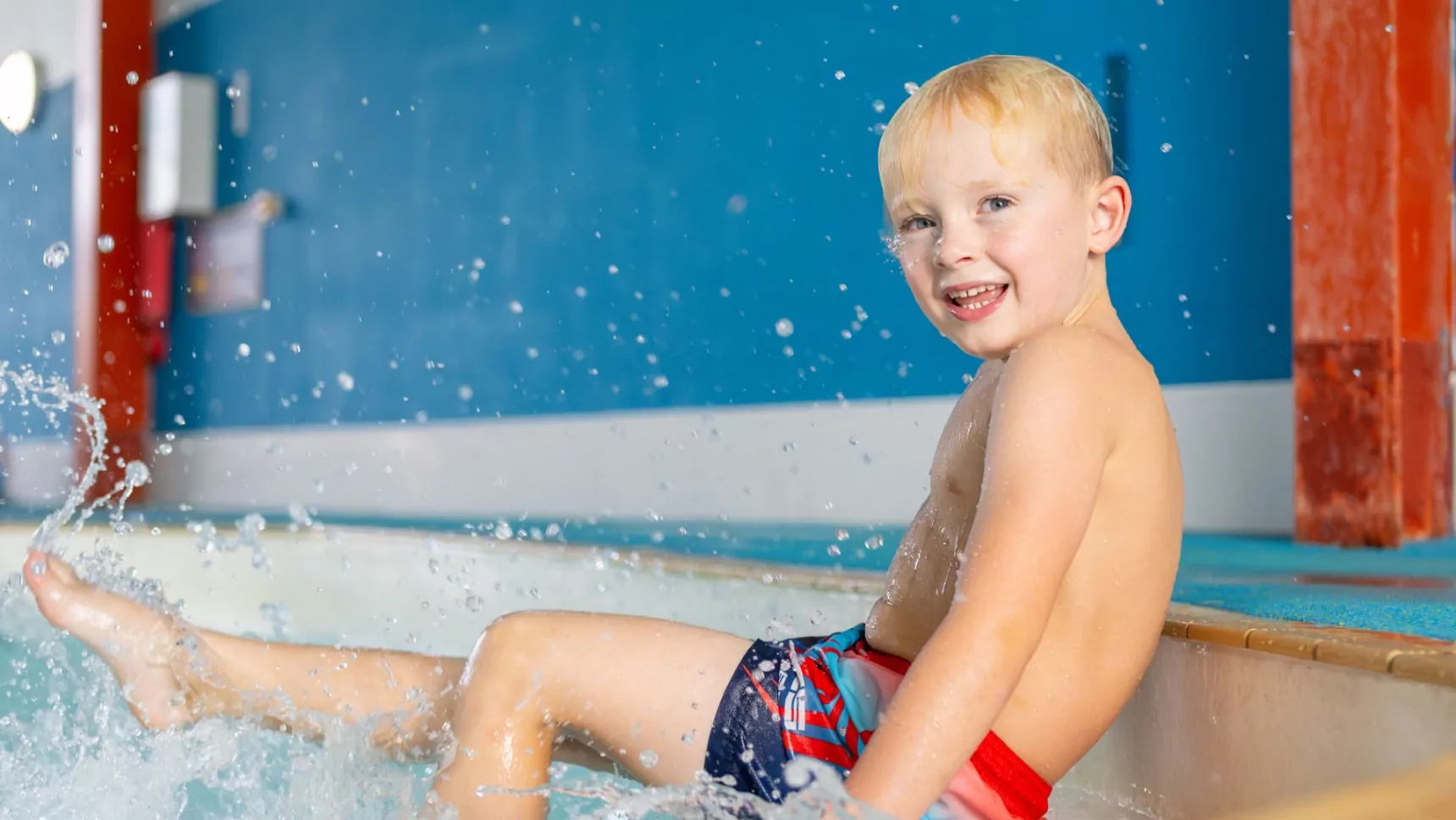 A smiling young boy splashing water while sitting on the edge of an indoor swimming pool, wearing colorful swim trunks, with a bright blue wall in the background.