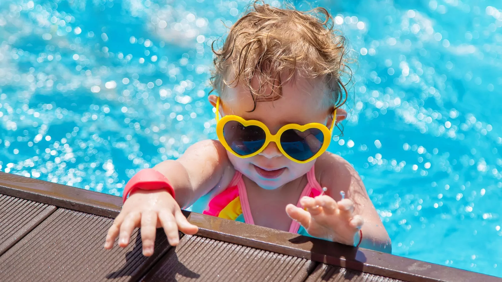 A young child wearing heart-shaped yellow sunglasses and a colorful swimsuit climbing out of a bright blue swimming pool, with wet hair and a playful expression.