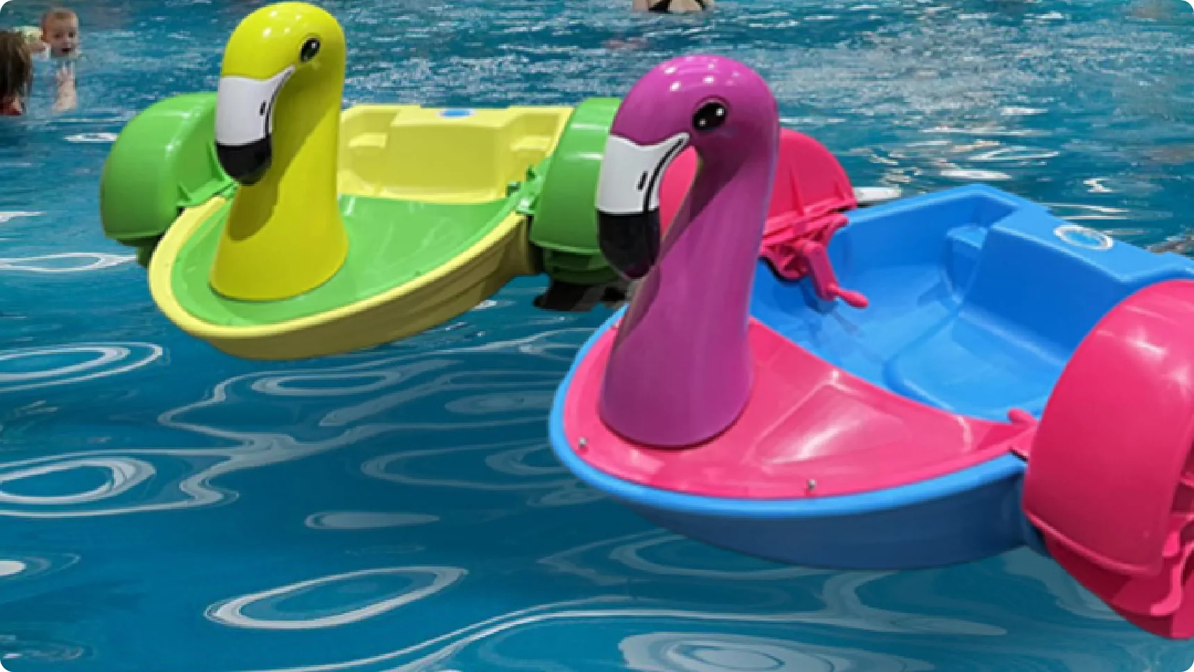 Two colorful paddle boats shaped like flamingos, one pink and one yellow, floating in an indoor swimming pool with children playing in the background.