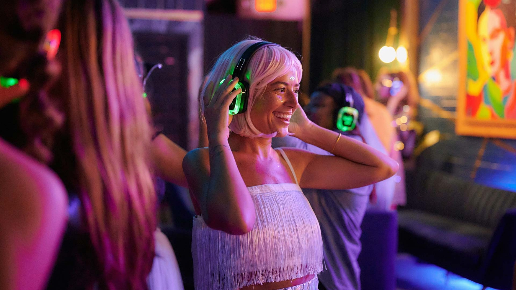 A woman in a white fringe outfit and pink wig enjoying a silent disco, wearing glowing green headphones, surrounded by people dancing in a vibrant, neon-lit party atmosphere.