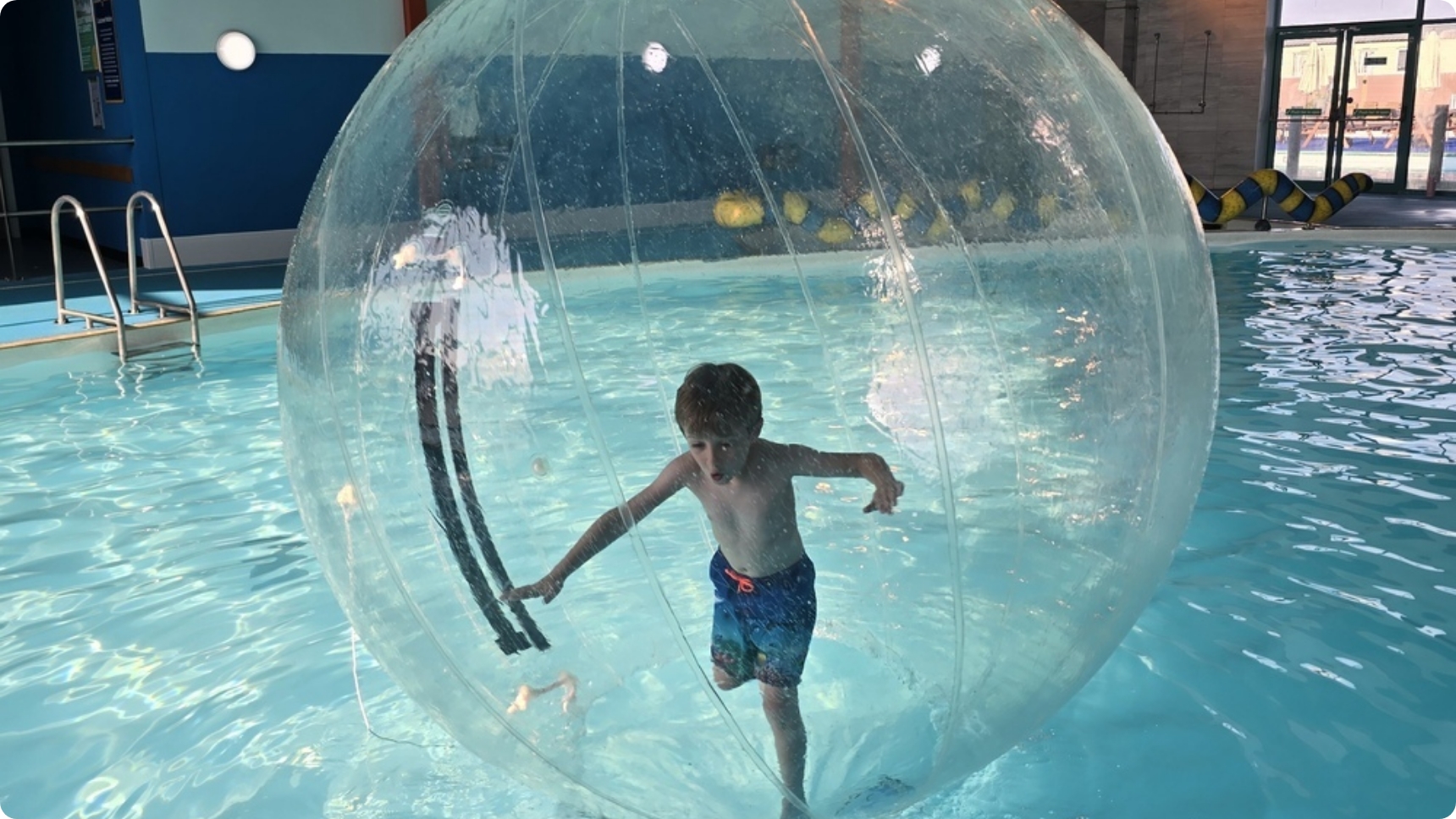 A young boy inside a giant inflatable water walking ball, balancing as he moves across the surface of an indoor swimming pool.
