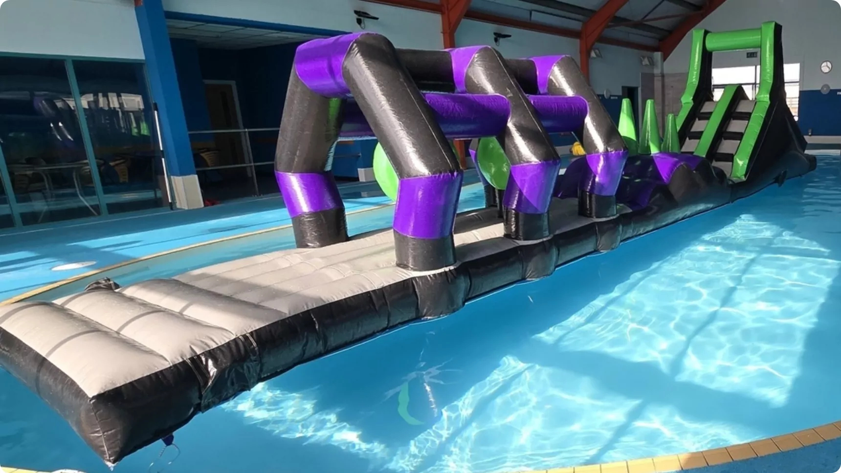 A large inflatable obstacle course floating in an indoor swimming pool, featuring black, purple, and green elements with climbing sections and a slide.