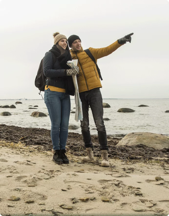 A couple dressed in warm clothing standing on a rocky beach, holding a map while the man points into the distance. They appear to be exploring the coastline on a cool day.
