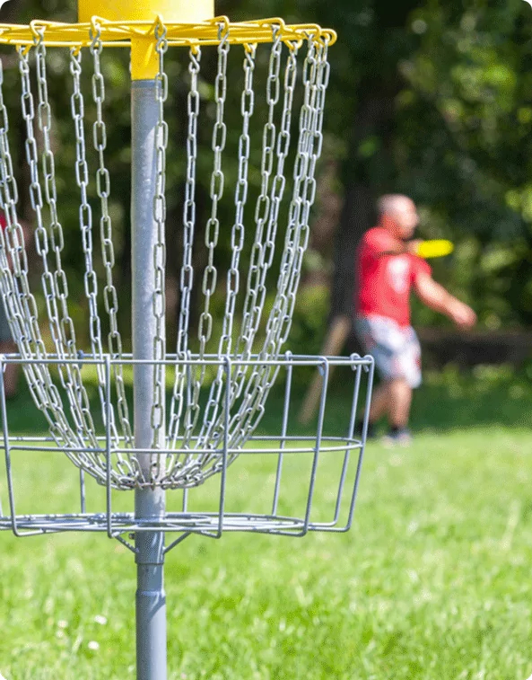 A close-up of a metal disc golf basket with chains, set on a grassy field. In the blurred background, a player in a red shirt is throwing a disc towards the target