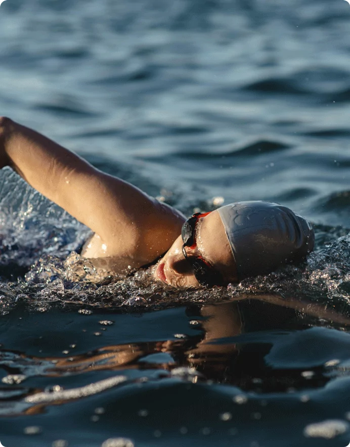 A swimmer wearing a silver swim cap and goggles performing a front crawl stroke in open water. The sunlight reflects off the rippling waves, creating a serene and athletic scene.
