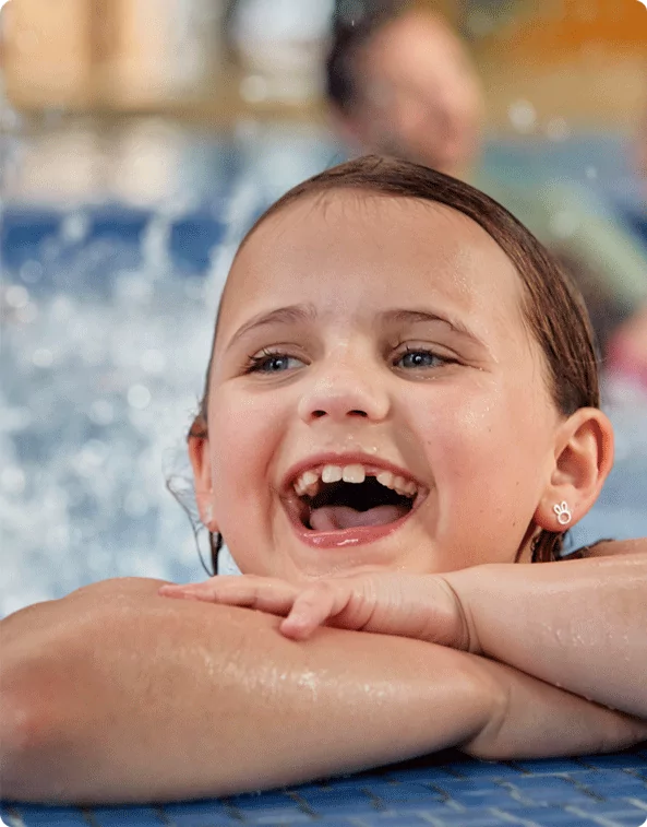 A young girl with wet hair and a big smile leaning on the edge of a swimming pool, laughing as water splashes behind her.