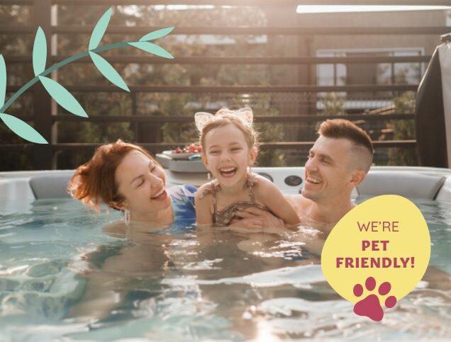 A smiling family enjoying a warm soak in a hot tub. A mother, father, and their young daughter, who is wearing cat-ear headbands, share a joyful moment together. A ‘We’re pet-friendly!’ sign is displayed in the corner.