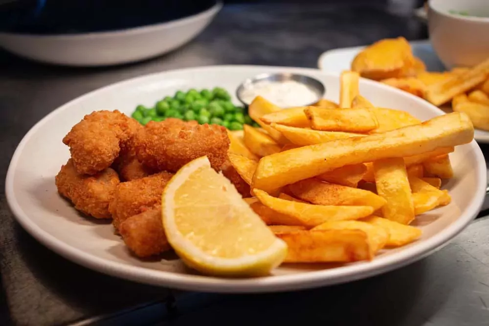 Plate of crispy fish fingers served with golden chips, green peas, a lemon wedge, and a pot of tartar sauce at Hayling Island.