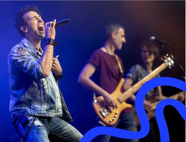 A passionate lead singer performing on stage, wearing a denim jacket and singing into a microphone. Behind him, a bass guitarist and an electric guitarist play under blue stage lighting.