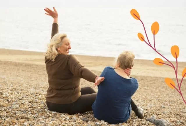Two older women sitting on a pebble beach by the sea, enjoying a relaxed and cheerful moment. One woman raises her arm in excitement while the other looks out at the ocean.