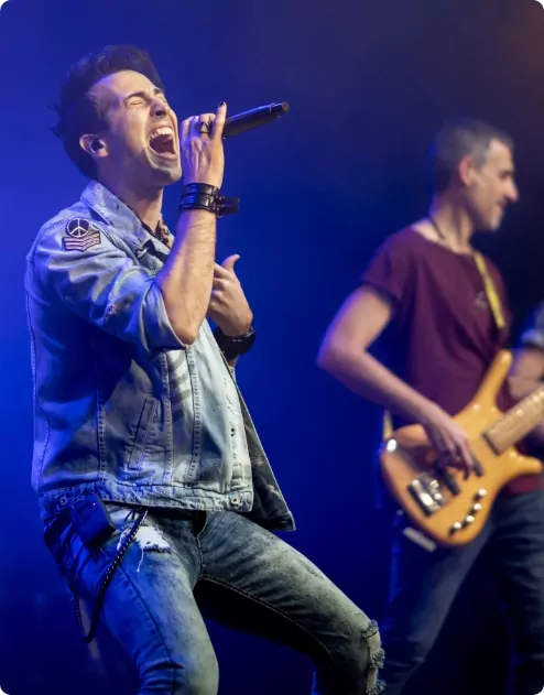 Man on stage singing into a microphone whilst his bandmate plays the guitar in the background