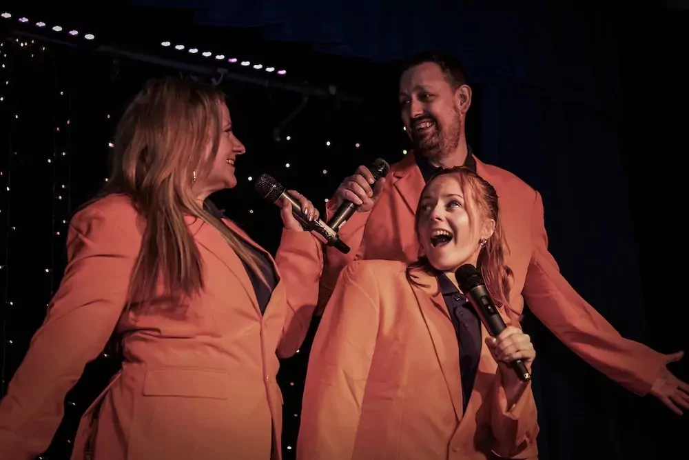 Three performers in matching orange jackets singing together on stage during a lively evening show at Hayling Island.