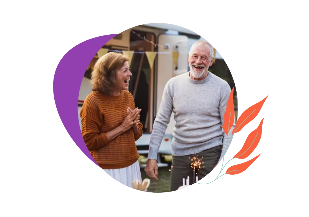 An older couple celebrating, the woman in a brown top clapping, and the man in a grey sweater holding a small cake with a sparkler, with a caravan in the background, Hayling Island.