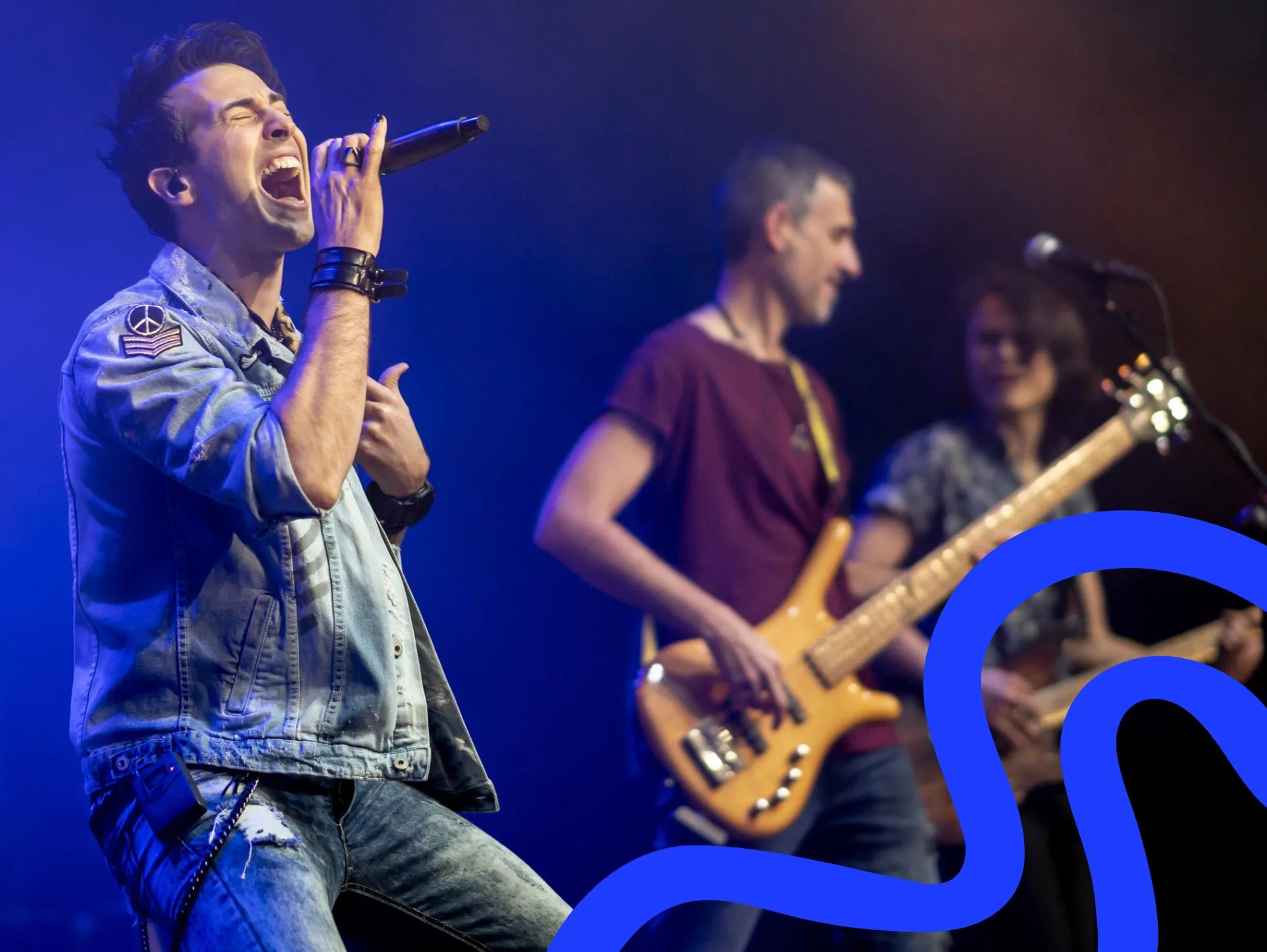 Alt Tag: A male singer enthusiastically performing on stage with a microphone, wearing a denim jacket, alongside a bassist and another musician in the background, Hayling Island.