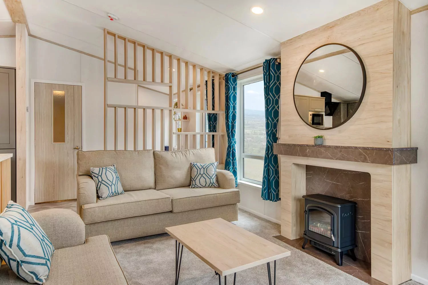 A modern and inviting living room in a holiday lodge, featuring two beige sofas, a light wood coffee table, a fireplace with a stove, and blue patterned curtains, Hayling Island.