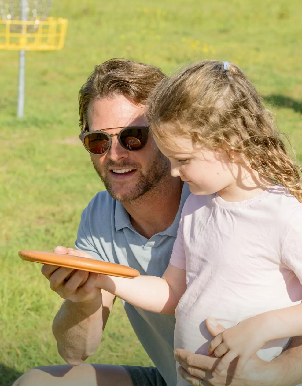 A father shows his young daughter how to hold a disc while playing disc golf on the sunny lawn at Hayling Island.