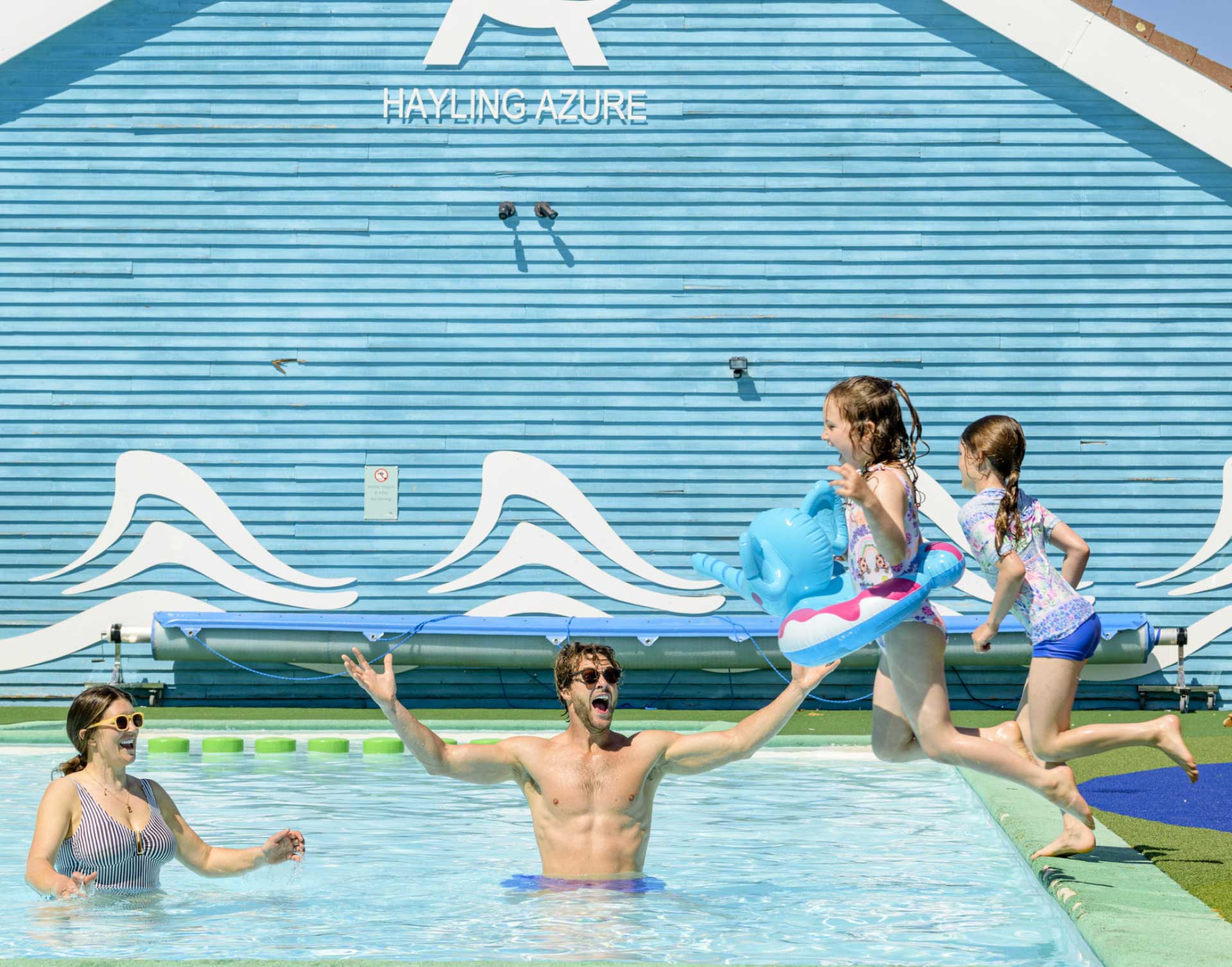 A family plays happily by the side of the outdoor pool, featuring the Hayling Azure building background at Hayling Island.