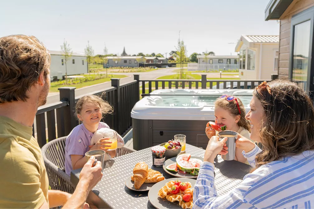 Family enjoying breakfast together on the decking of a holiday lodge with a private hot tub in the background at Hayling Island