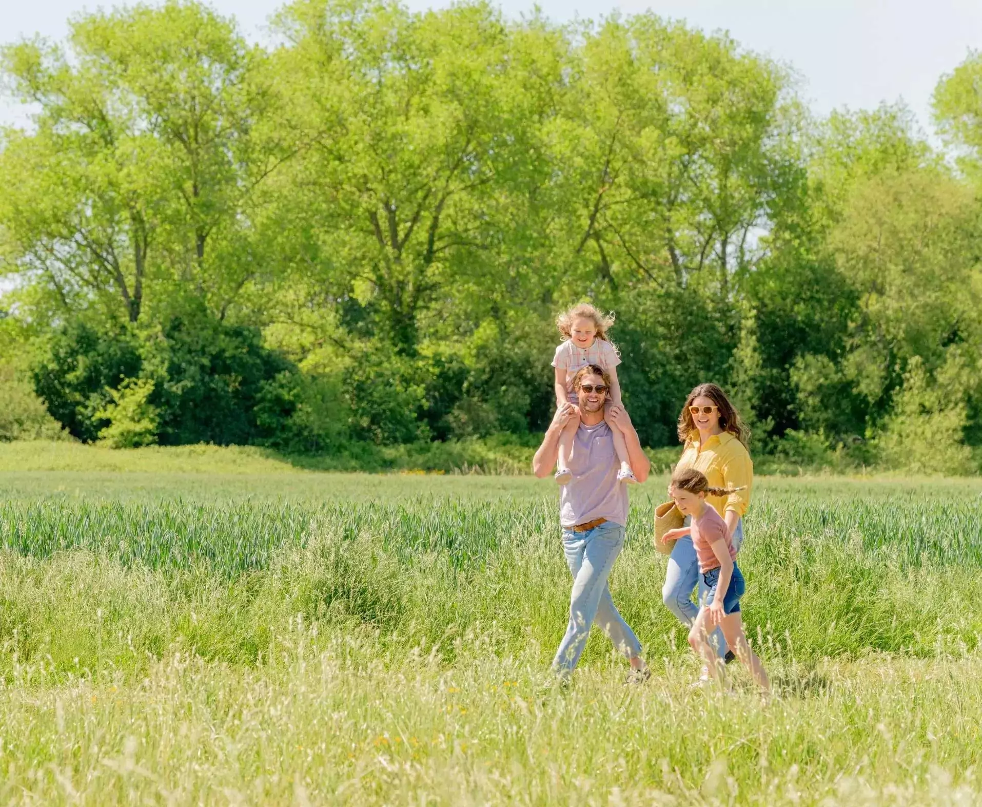 A happy family walking through a sunny countryside field, with a father carrying his young daughter on his shoulders at Hayling Island.
