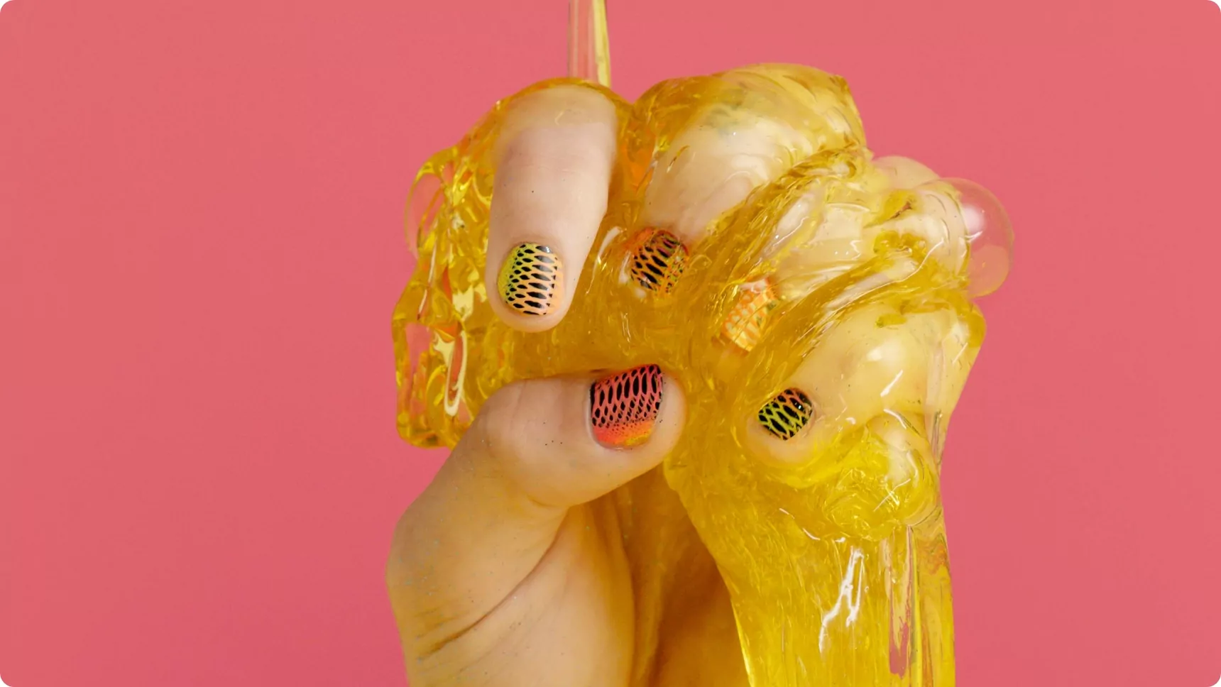 A hand squeezing bright yellow slime against a bold pink background, with nails painted in a black and orange gradient design featuring a mesh pattern.