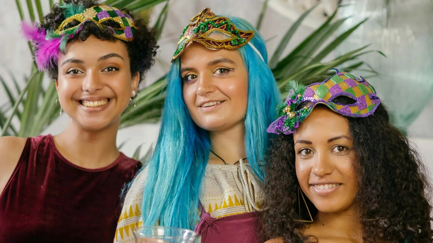 Three smiling women dressed in festive attire wearing colorful masquerade masks, with one sporting a vibrant blue wig, celebrating at a lively party.
