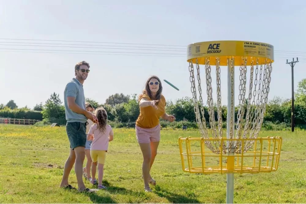 Family playing disc golf together on a sunny day at Hayling Island, aiming for the yellow target basket.