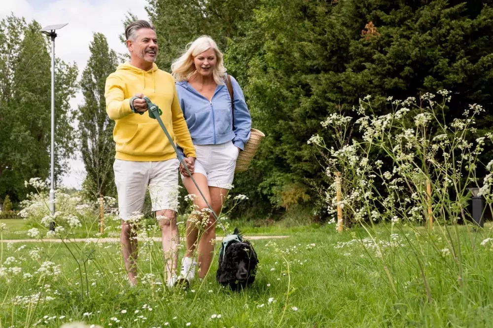 A smiling couple, the man in a yellow hoodie holding a leash with a black dog, and the woman in a blue hoodie, walking together through a grassy area with trees, Hayling Island.