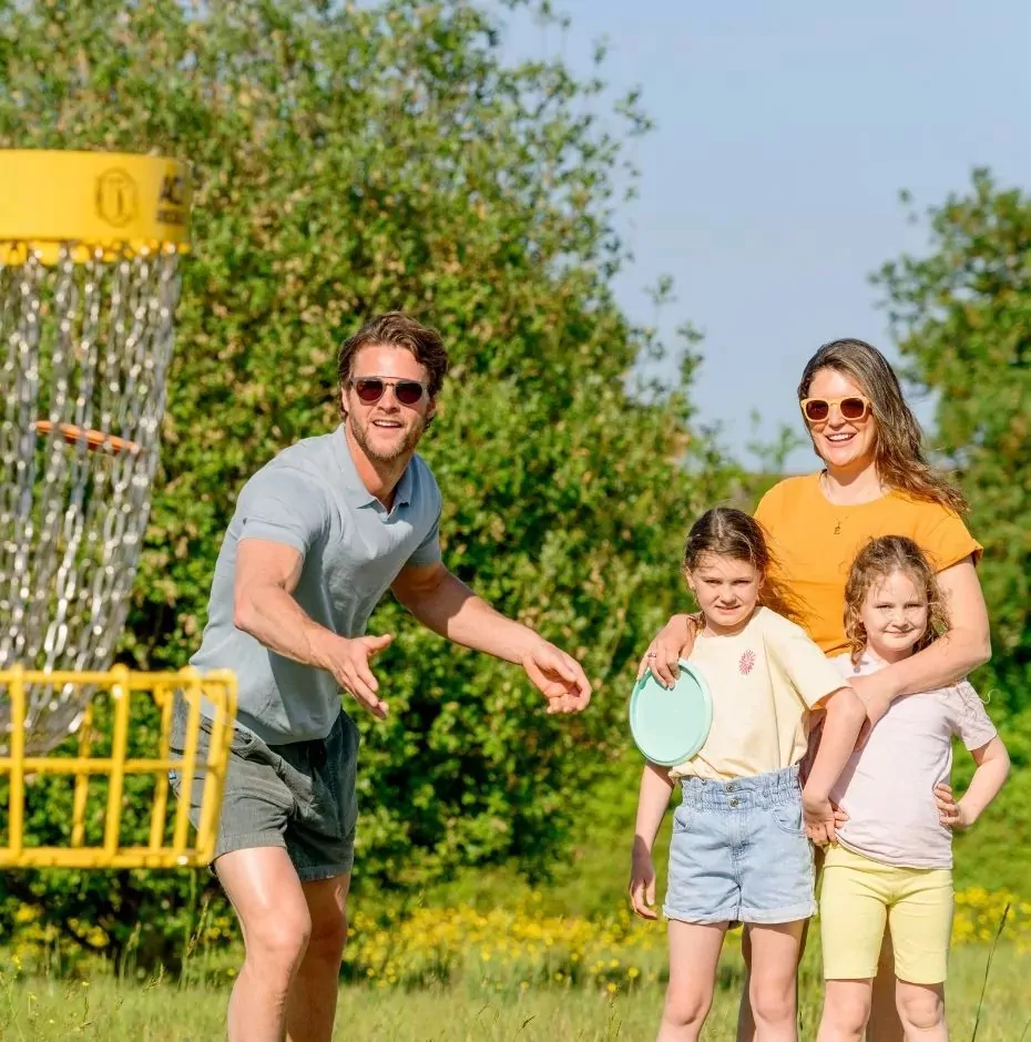 A happy family playing disc golf on a sunny day, with a man throwing a disc towards a yellow basket and a woman with two young girls watching, Hayling Island.
