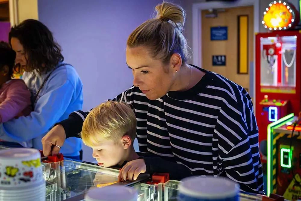 A mother and her young son focused on playing an arcade game, with other arcade machines and people in the background, Hayling Island.