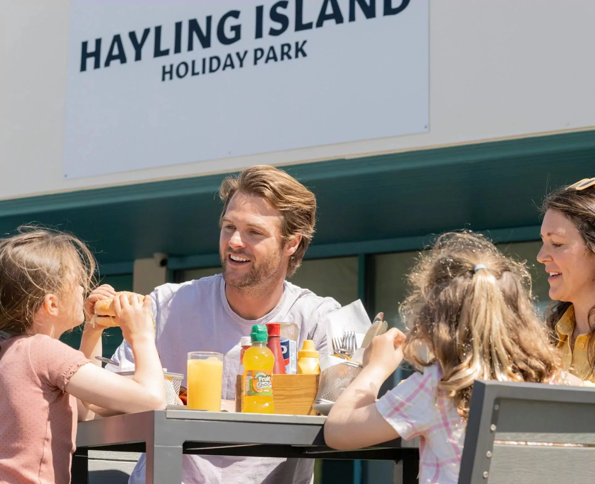 A smiling family of four, including two adults and two young girls, enjoying an outdoor meal at a table with drinks and food, under a sign for "Hayling Island Holiday Park", Hayling Island.
