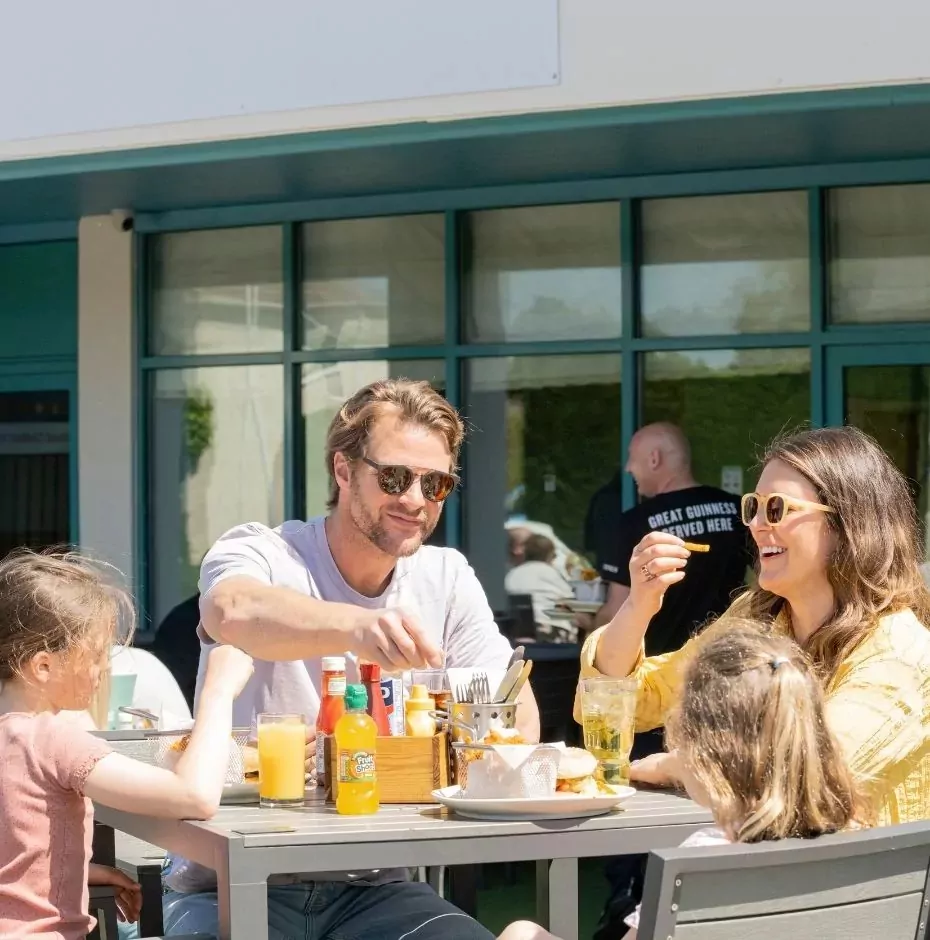 A family of four enjoying an outdoor meal at a restaurant patio, with a man, woman, and two young girls laughing and eating in the sunshine, Hayling Island.
