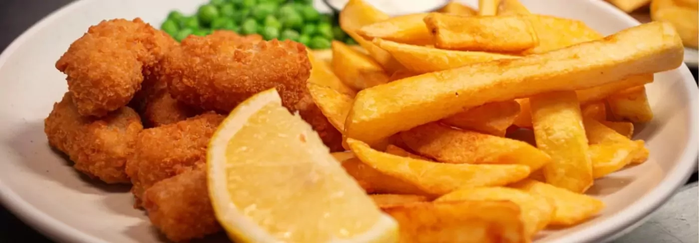 A close-up plate of golden-brown fish nuggets, thick-cut chips, a wedge of lemon, and green peas, representing a classic meal available, Hayling Island.