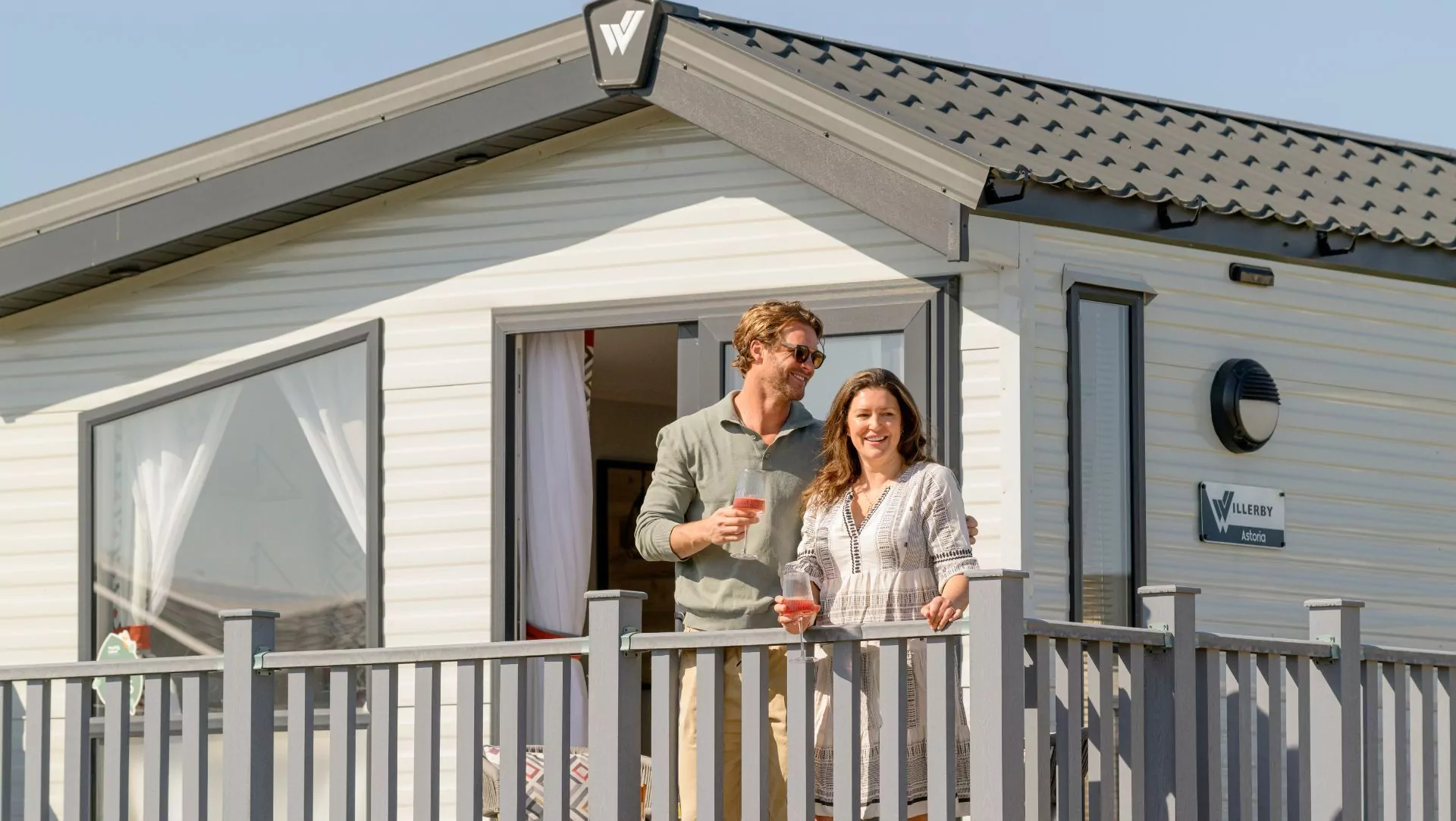 A happy couple, a man and a woman, standing on the balcony of a modern, light-colored holiday home, each holding a pink drink, enjoying the sunny weather, Hayling Island