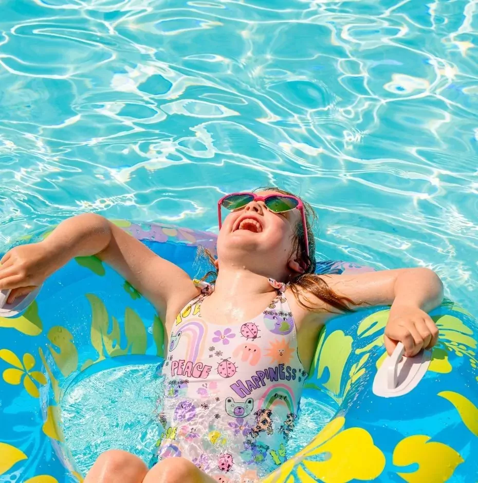 A young girl in pink sunglasses and a colorful swimsuit laughing joyfully while floating in a bright blue swimming pool on an inflatable ring, Hayling Island.