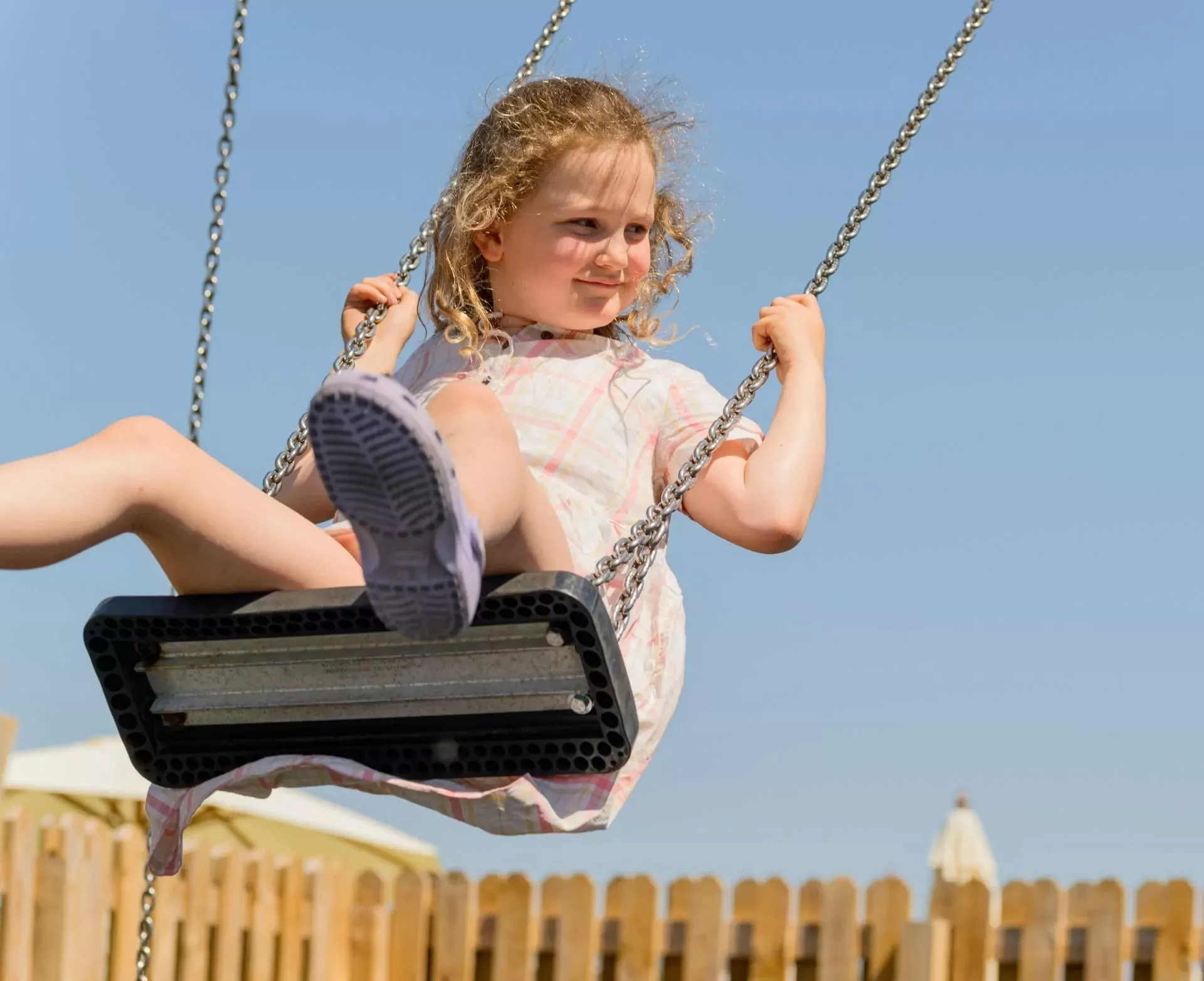 A joyful young girl with curly hair swinging high on a playground swing under a clear blue sky, smiling and looking to the side, Hayling Island.