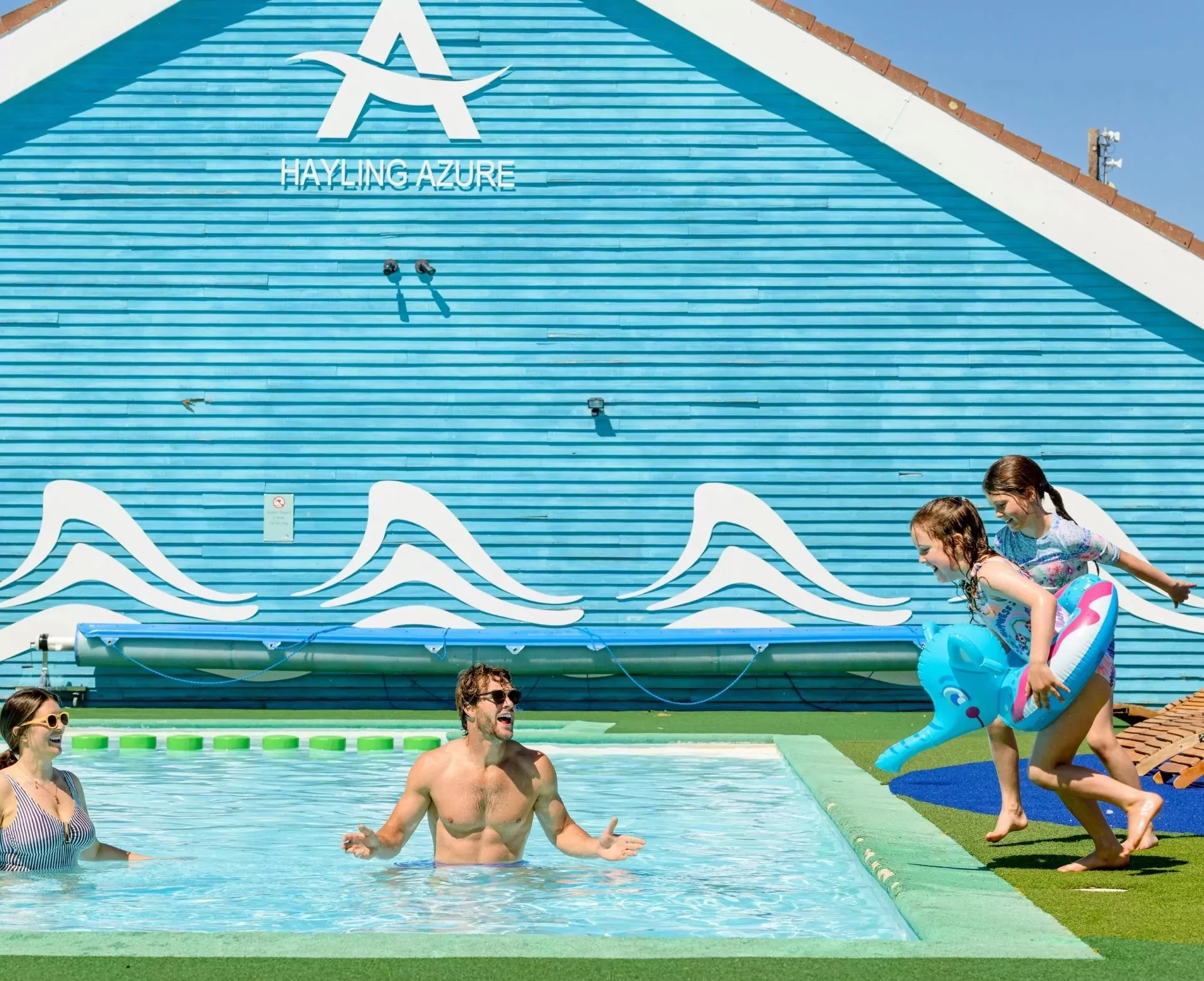 A family enjoying a swimming pool at Hayling Azure, with a man in the water and two young girls running with inflatables by the poolside, and a blue building in the background, Hayling Island.