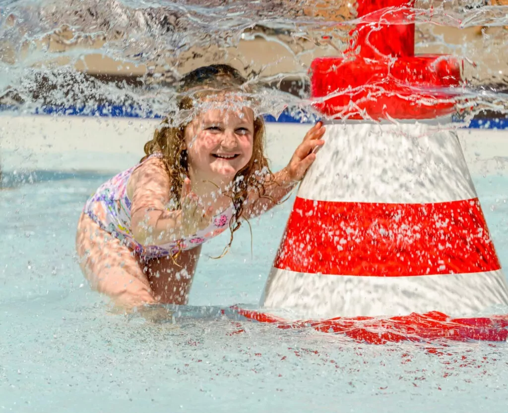 A happy young girl with curly hair laughing and splashing in a shallow pool next to a red and white striped water feature, with water spraying around her, Hayling Island.