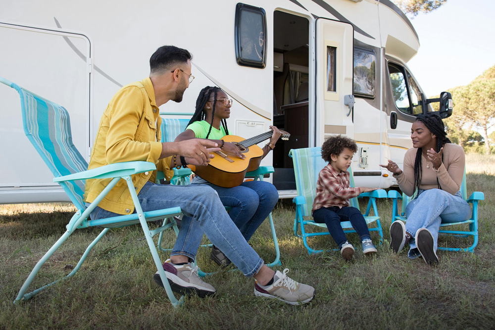 A happy family playing guitar and relaxing in chairs outside their motorhome at Henfold Lakes.