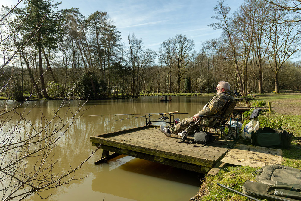 A man fishing from a wooden platform on a calm lake surrounded by trees during a stay at Henfold Lakes.