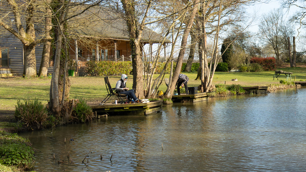 Two anglers fishing from wooden pegs on the tree-lined lake bank at Henfold Lakes, with a large lodge building visible behind.