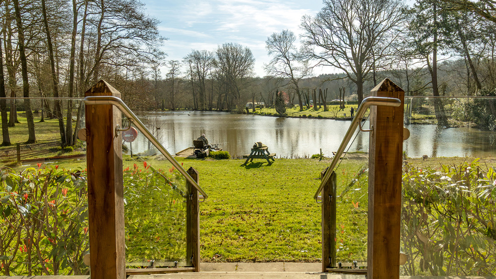 Looking through a glass and wood balustrade entrance over a grassy bank towards the main fishing lake at Henfold Lakes.