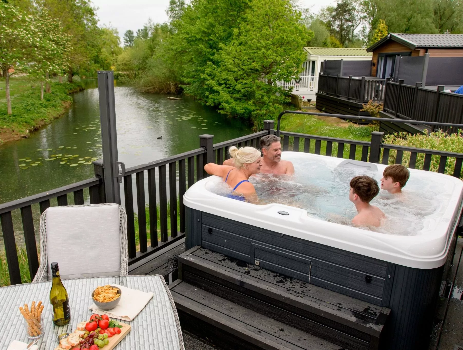 Family enjoying a hot tub on a wooden deck beside a river, with a picnic table set with food and drinks, promoting luxury lodge living at Meadow Bay Villages.