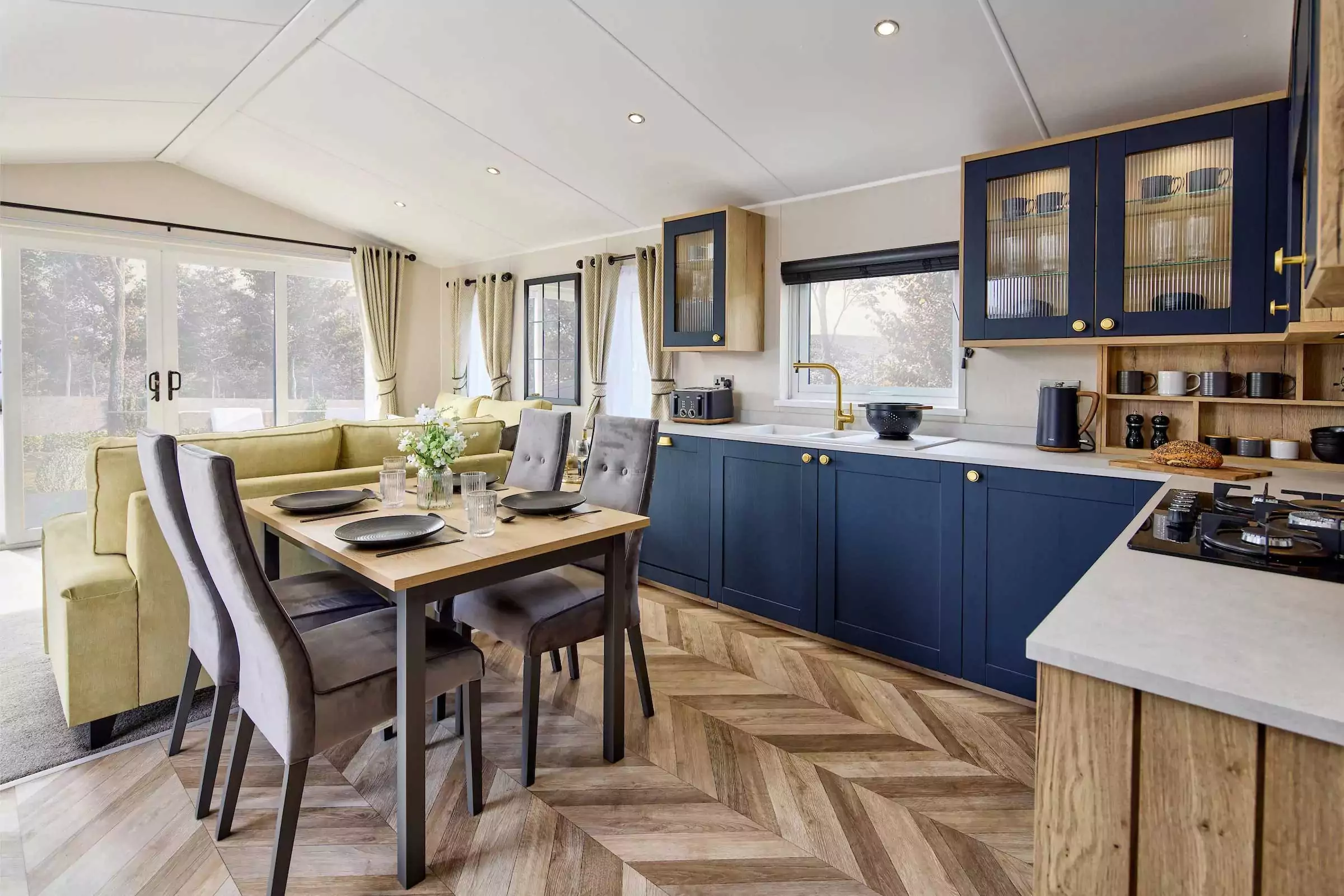 Stylish open-plan kitchen and dining area inside a platinum-grade caravan at Meadow Bay Villages, with navy cabinets, chevron flooring, and modern decor.