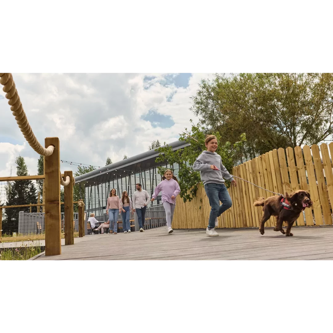 Children running with a dog on a lead along a wooden boardwalk, followed by smiling family members outside a modern glass-fronted holiday park building.