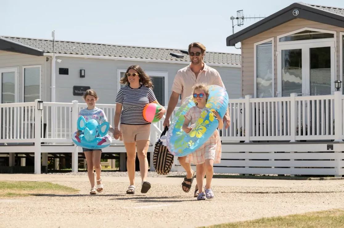 Smiling family walking with colourful pool floats and beach toys outside their Meadow Bay Villages holiday lodge.