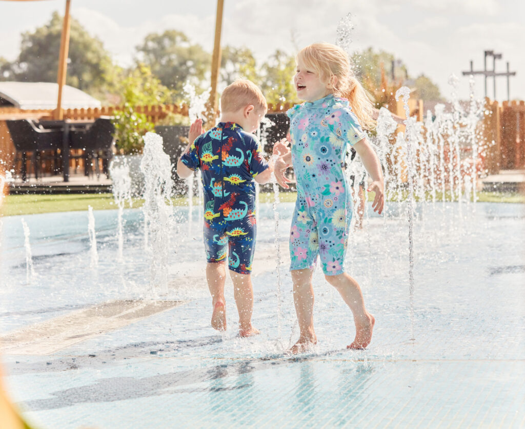 Young children playing and laughing in a splash pad water feature, wearing colourful swimsuits on a sunny day at the holiday park.