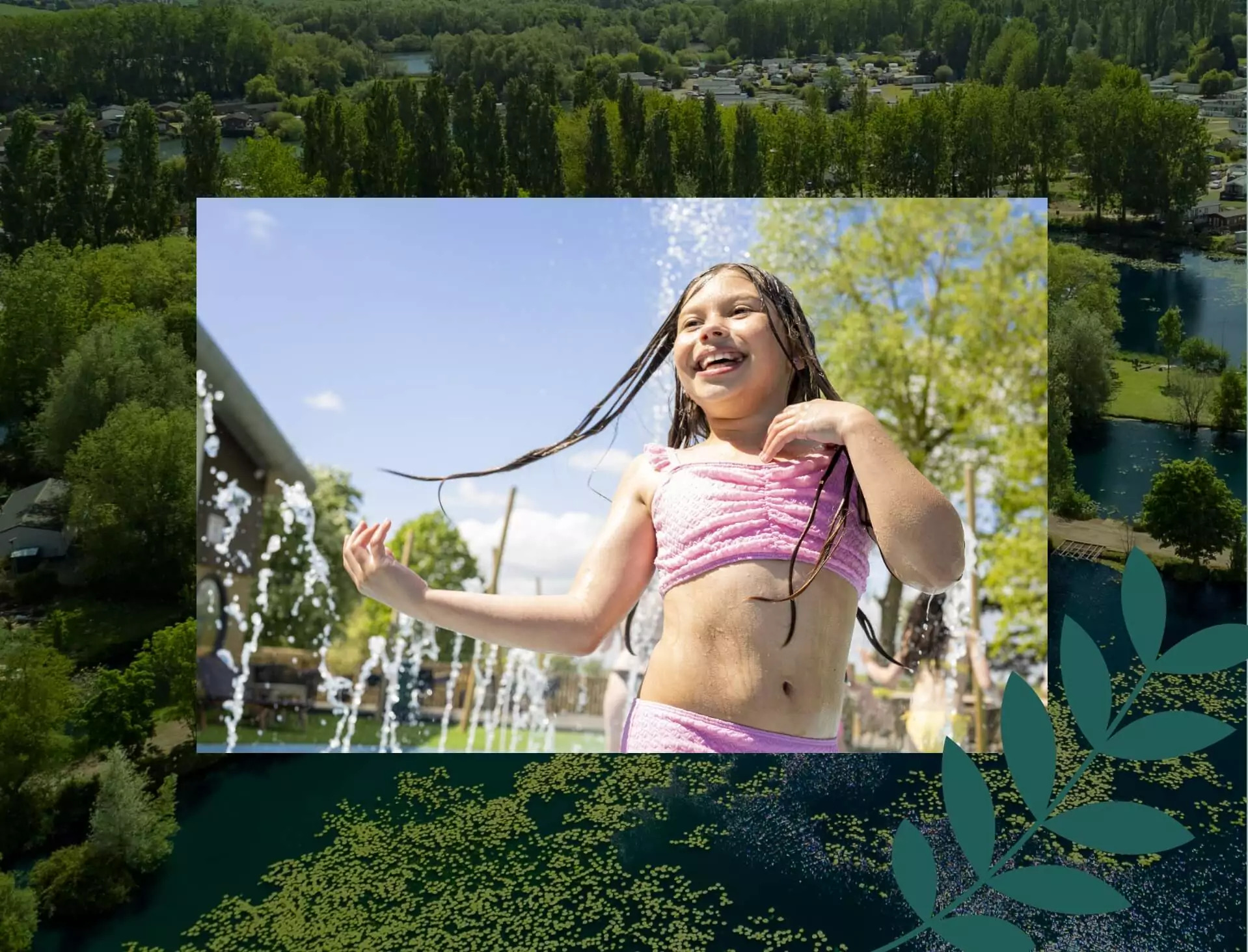 Smiling young girl playing in an outdoor splash zone water feature, wearing a pink swimsuit on a sunny day at the holiday park.